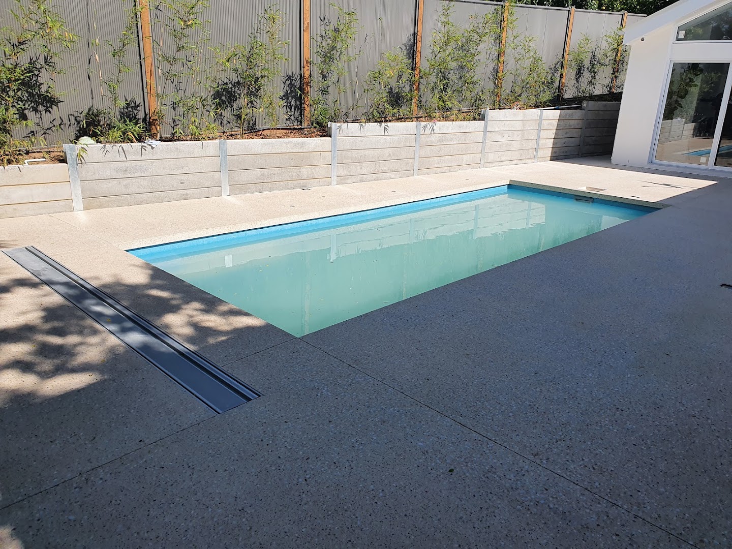 A pool with a concrete surround beside a rendered retaining wall and garden