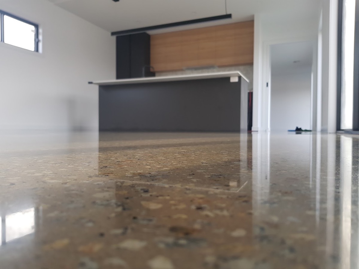 A polished concrete floor with exposed stone reflecting a kitchen bench and joinery