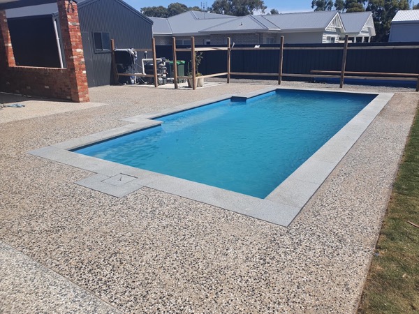 A pool with an exposed aggregate concrete surround beside a dark fence